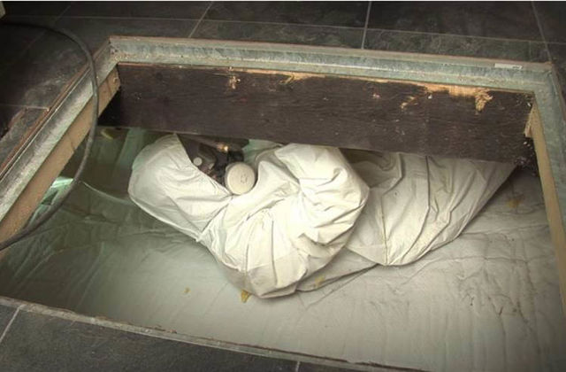 Man in witte beschermingsoverall in de kruipruimte is bezig vloerisolatie aan te brengen.