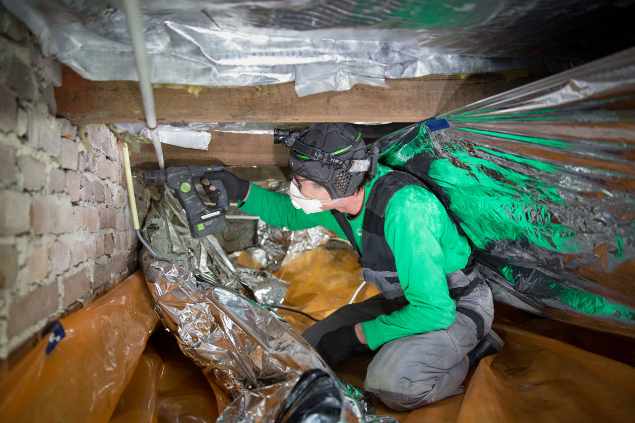 Een man maakt isolatiemateriaal, in dit geval isolatiekussens, vast in de kruipruimte onder de vloer. De man draagt beschermende kleding.