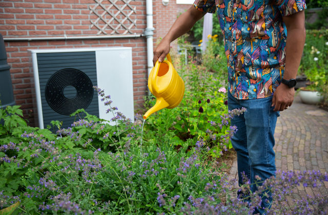 Buitenunit van een warmtepomp in tuin tegen de muur. Ervoor staan bloeiende planten, man geeft water.