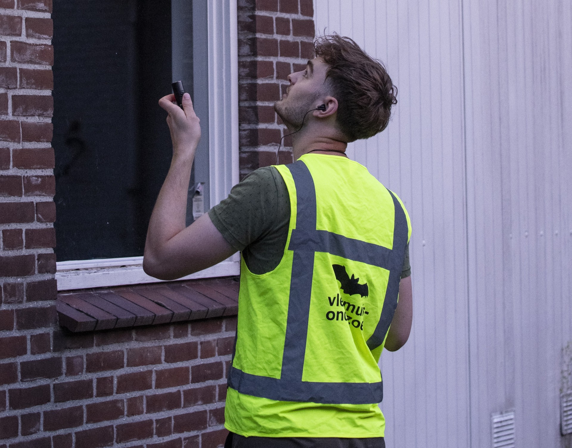 Een ondersteuner van een ecologisch bureau met een geel hesje staat naast de gevel van een gebouw met een scanner voor vleermuizen.