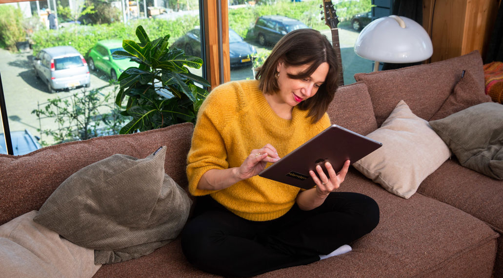 Een vrouw zit met haar tablet op de bank van haar appartement.