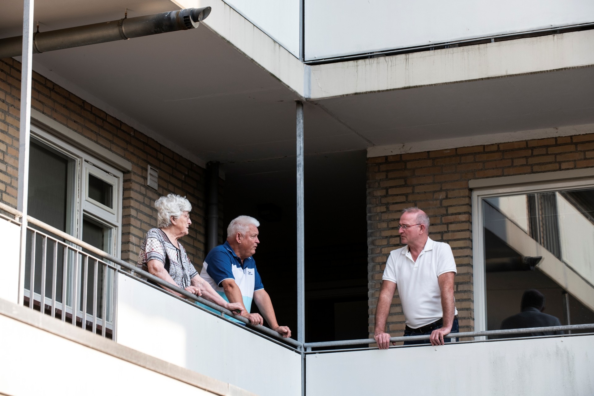 Twee oudere mensen zijn op de galerij van een flat in gesprek met een man in een wit shirt.