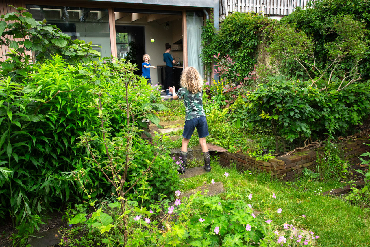 Een foto van een jongen met krullend haar gooit een bal over met een blonde jongen in een groene tuin vol planten.