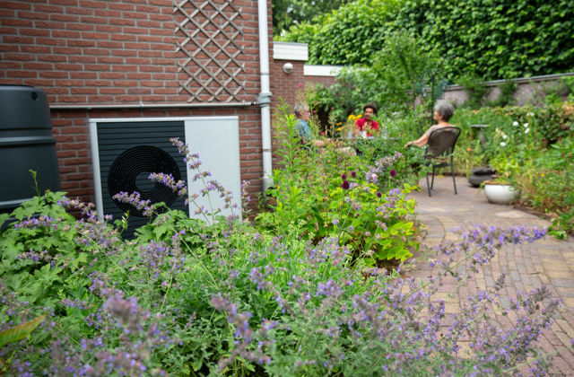 Buitenunit van warmtepomp in tuin. Op de voorgrond een bloeiende plant, op achtergrond mensen op terras.