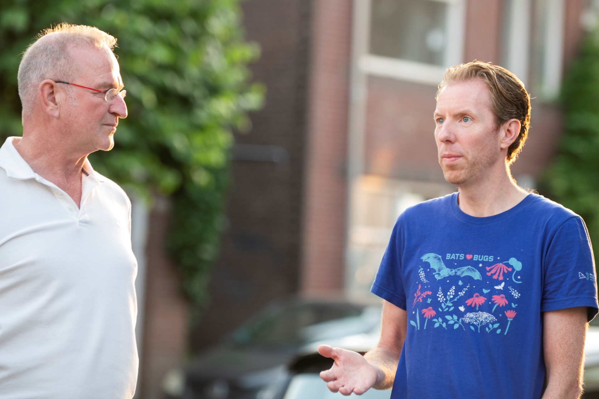 Twee mannen zijn in gesprek. Eén man draagt een wit shirt en een bril en heeft grijzend haar. De andere man heeft rossig halflang haar en draagt een blauw shirt met de tekst Bats love Bugs.