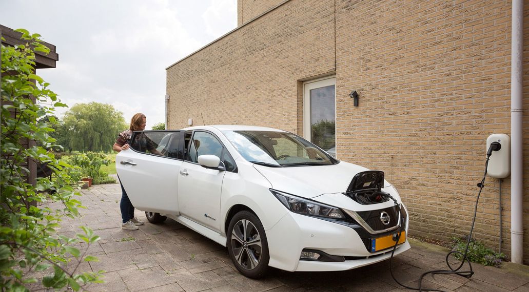 Op de foto staat een witte elektrische auto op een oprit naast een huis. De auto is met een snoer is verbonden aan de laadpaal. De laadpaal hangt aan de zijgevel van de woning. Een vrouw staat naast de auto en pakt iets van de achterbank. 