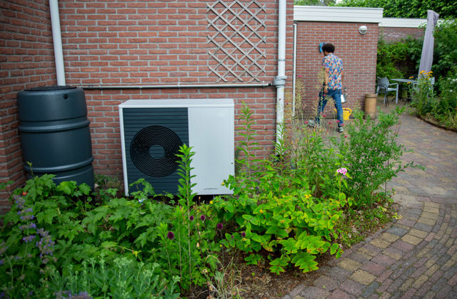 Buitenunit van een warmtepomp tegen huis. Ernaast een regenton, op de achtergrond loopt een man.